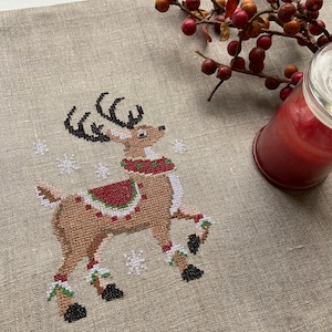 May include: A cross-stitch design of a reindeer with a red and green saddle on a beige linen fabric background. The reindeer is wearing a red and green scarf and has black hooves. There are white snowflakes in the background.