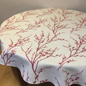 May include: A round tablecloth with a cream background and a repeating pattern of red and white flowering branches. The tablecloth is draped over a dark wooden table, showcasing the floral design.