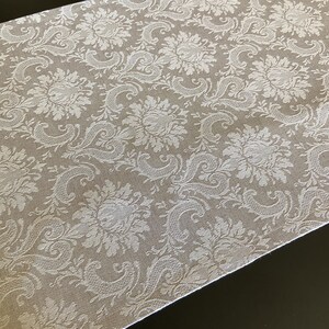 Peut inclure: Chemin de table en damas beige et blanc. Le motif répété présente des fleurs et des volutes. Le tissu a un léger éclat et les bords sont soigneusement finis. Le chemin de table est disposé en diagonale sur une surface sombre.