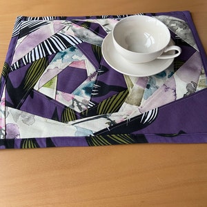 May include: A purple placemat with a geometric pattern of black, white, green, and pink shapes. A white teacup and saucer are on top of the placemat.