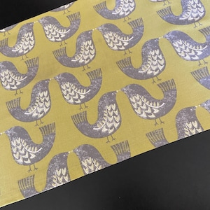 Puede incluir: Una tela decorativa con un patrón repetido de pájaros grises estilizados sobre un fondo amarillo-verdoso. Los pájaros se enfrentan, con detalles blancos en sus alas y cuerpos. La tela tiene una textura suave y está en ángulo en el marco.