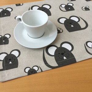 May include: A gray and white linen placemat with a repeating pattern of black mice. A white cup and saucer are on top of the placemat.
