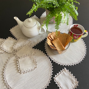 Puede incluir: Un juego de seis manteles individuales de lino beige con un delicado borde de encaje. Los manteles individuales están dispuestos sobre una superficie negra con una tetera blanca, un cuenco de galletas y una taza de té.