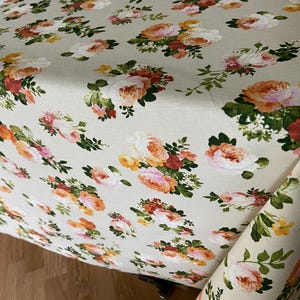 Può includere: Una tovaglia in tessuto beige chiaro caratterizzata da un motivo floreale ripetuto di rose e piccoli fiori nelle tonalità dell'arancione, pesca, rosa e bianco, arricchito da foglie e steli verdi distribuiti sull'intera superficie del tessuto.