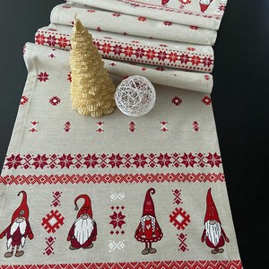 Könnte beinhalten: Ein beiger Tischläufer mit einem rot-weißen skandinavischen Muster. Der Läufer zeigt ein sich wiederholendes Muster aus Schneeflocken und Gnomen. Der Läufer ist in der Hälfte gefaltet, wobei die obere Schicht einen Gnom mit roter Mütze und Bart zeigt.