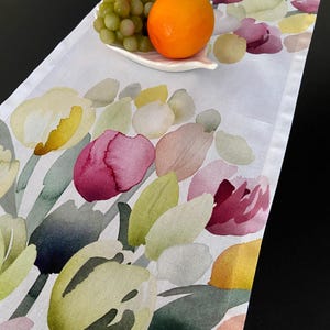 Può includere: Runner da tavolo bianco con un design floreale ad acquerello con forme di tulipani gialli, rosa e verdi. Un piccolo piatto bianco contiene uva, un'arancia e un piccolo frutto. Il runner è su una superficie nera.