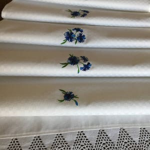 Op de afbeelding: Witte linnen tafelloper met geborduurde blauwe korenbloemen. De loper heeft een geweven textuur en een decoratieve gehaakte kanten rand. De korenbloem borduursels zijn in verschillende maten en opstellingen.
