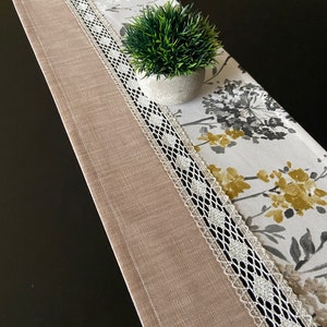 Puede incluir: Un camino de mesa beige con un borde de encaje blanco y un patrón floral. Una pequeña planta en maceta se encuentra sobre el camino de mesa.