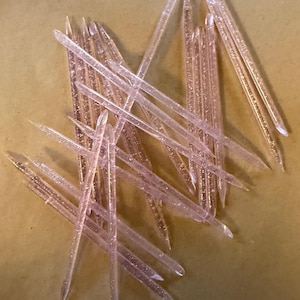 May include: A collection of translucent pink plastic sticks with glitter, scattered on a light brown surface. The sticks are pointed at one end and appear to be decorative or craft supplies.