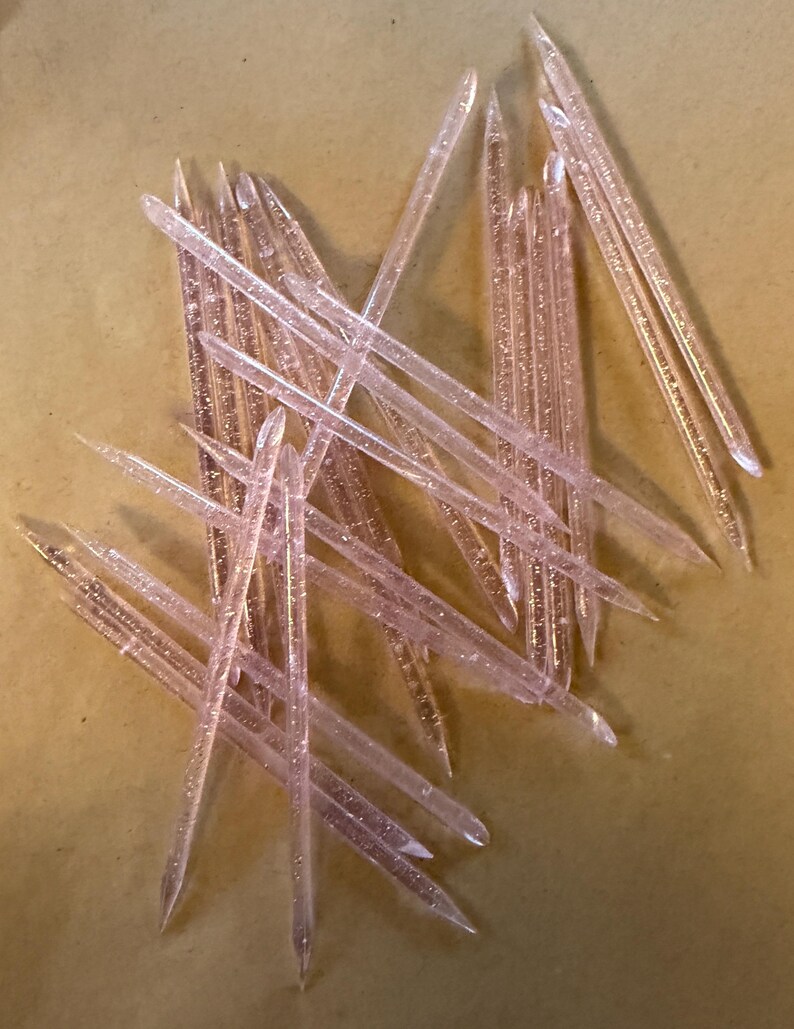 May include: A collection of translucent pink plastic sticks with glitter, scattered on a light brown surface. The sticks are pointed at one end and appear to be decorative or craft supplies.