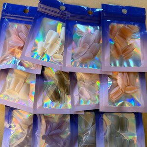 May include: Assortment of false nail sets in various colors, including pink, white, and gray. Each set is packaged in a clear, iridescent pouch with a blue top. The nails are in different shapes, such as oval and square.