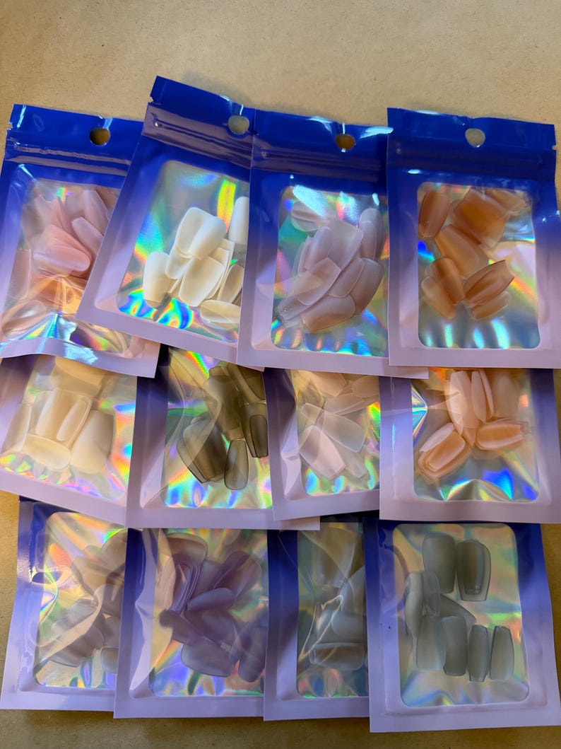May include: Assortment of false nail sets in various colors, including pink, white, and gray. Each set is packaged in a clear, iridescent pouch with a blue top. The nails are in different shapes, such as oval and square.