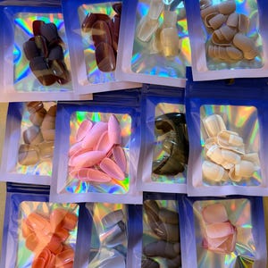 May include: A collection of various artificial nail sets in clear plastic bags with blue zip closures. The nails come in a variety of colors, including pink, nude, and brown. The bags have a holographic iridescent effect.