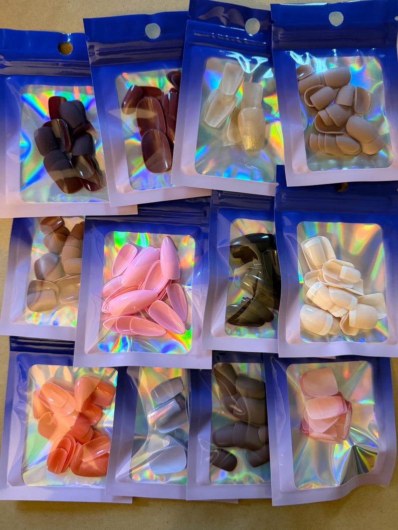 May include: A collection of various artificial nail sets in clear plastic bags with blue zip closures. The nails come in a variety of colors, including pink, nude, and brown. The bags have a holographic iridescent effect.