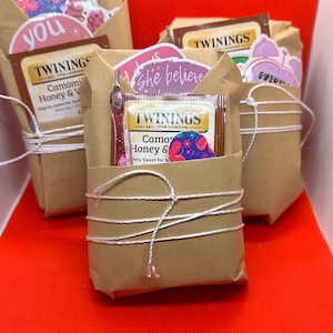 May include: Three wrapped gift sets on a red surface. Each set is wrapped in brown paper and tied with white string. The sets contain tea bags, stickers with text, and a small pink item. The tea bags are from Twinings.