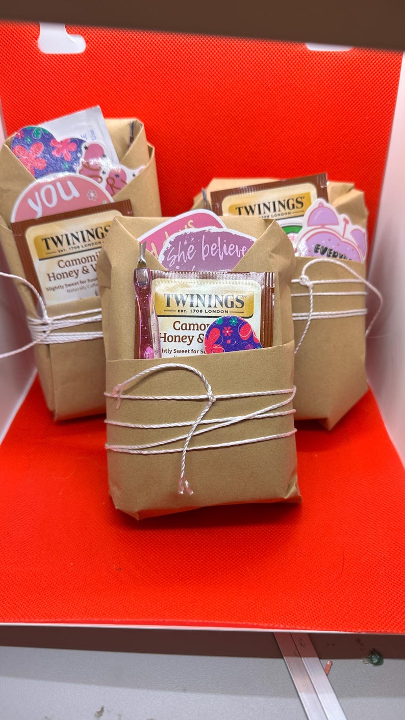 May include: Three wrapped gift sets on a red surface. Each set is wrapped in brown paper and tied with white string. The sets contain tea bags, stickers with text, and a small pink item. The tea bags are from Twinings.