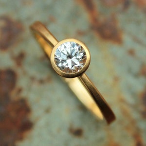 May include: A gold ring with a single round, clear diamond set in a bezel setting.
