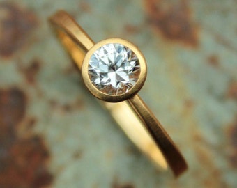 May include: A gold ring with a single round, clear diamond set in a bezel setting.