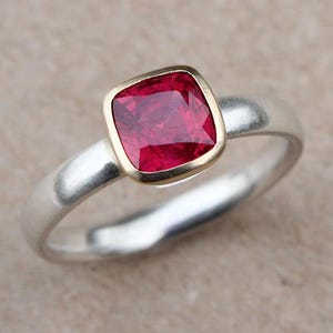 Könnte beinhalten: Ein silberner Ring mit einem quadratischen, leuchtend roten Edelstein, der in einer goldenen Lünette gefasst ist. Der Ring hat ein glattes, poliertes Band und ein klassisches Design. Der Edelstein misst etwa 0,8 cm.