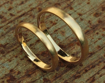 May include: Two gold wedding bands, one slightly wider than the other, are placed side-by-side on a rustic, brown, textured surface.