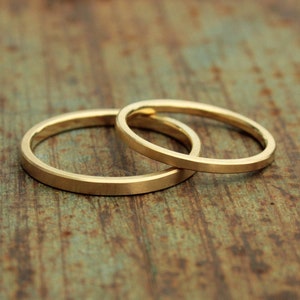 May include: Two gold wedding bands, one slightly larger than the other, are stacked on top of each other on a rustic wooden surface.