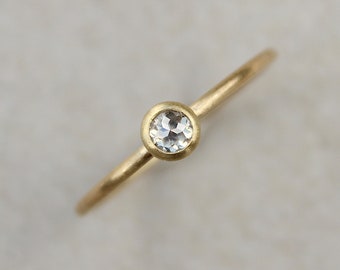 May include: A delicate gold ring featuring a small, round, clear gemstone set in a bezel. The ring has a thin band and a simple, elegant design. The gemstone is approximately 0.25 inches in diameter.