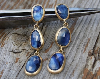 Puede incluir: Un par de pendientes de oro con tres piedras de zafiro azul cada uno. Los pendientes son colgantes y tienen un diseño delicado.