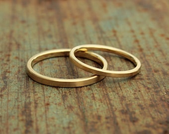 May include: Two gold wedding bands, one slightly larger than the other, are stacked on top of each other on a rustic wooden surface.