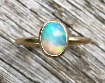 May include: A gold ring with an oval-shaped opal gemstone. The opal is a light blue color with flashes of green and orange.