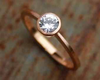 May include: A rose gold ring with a bezel setting featuring a round, clear gemstone.