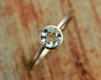 May include: A delicate ring with a round, light blue gemstone set in a thin, gold band. The ring is centered on a textured, brown surface, showcasing its simple elegance. The gemstone is approximately 0.25 inches in diameter.