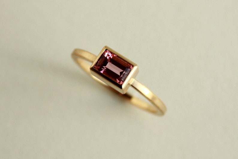 May include: A gold ring with a pink rectangular gemstone set in a gold bezel.