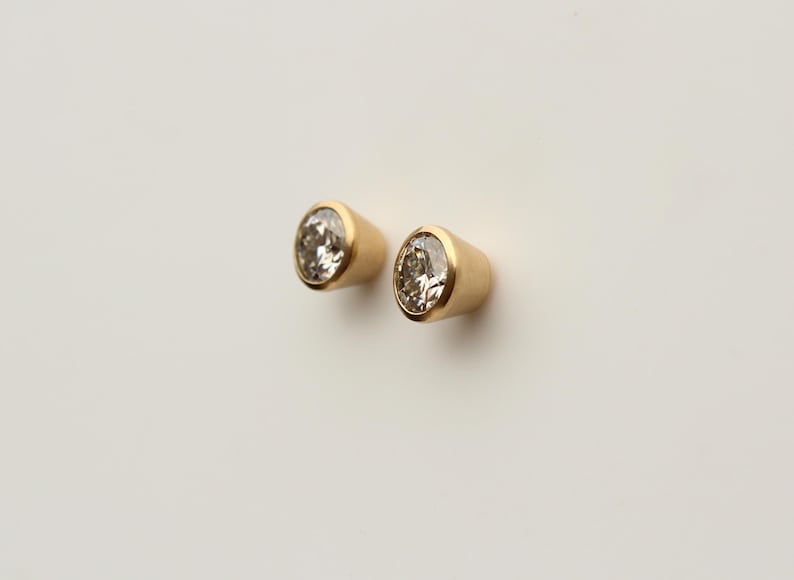 Puede incluir: Un par de pendientes de bot&oacute;n dorados. Cada pendiente presenta una piedra preciosa redonda y transparente engastada en un bisel dorado. Dise&ntilde;o sencillo y elegante, adecuado para el uso diario o ocasiones especiales.