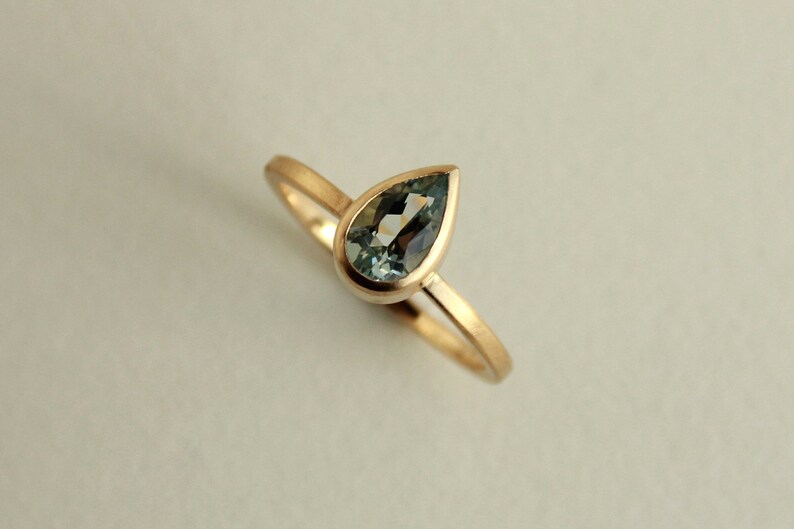 May include: A delicate gold ring with a pear-shaped aquamarine gemstone set in a bezel setting.