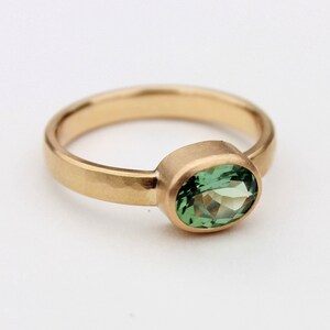 May include: A gold ring with a green oval gemstone set in a bezel setting. The band is hammered and has a matte finish.