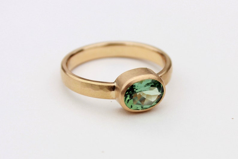 May include: A gold ring with a green oval gemstone set in a bezel setting. The band is hammered and has a matte finish.