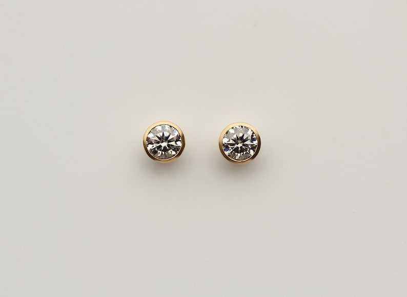 Puede incluir: Dos pendientes de diamantes redondos con engaste de bisel dorado. Los diamantes brillan sobre un fondo blanco. Estos pendientes cl&aacute;sicos son un elemento b&aacute;sico de joyer&iacute;a atemporal.