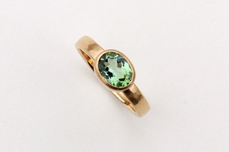 May include: A gold ring with a green oval gemstone set in a bezel setting. The band of the ring is hammered.