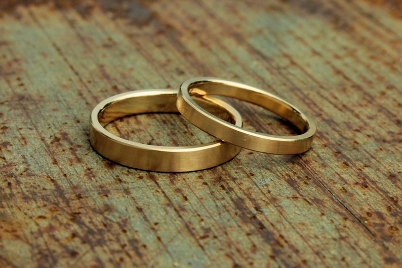 May include: Two gold wedding bands, one slightly larger than the other, are lying side by side on a rustic wooden surface.