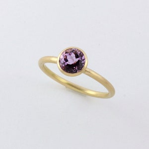 May include: A delicate gold ring with a round, bezel-set purple gemstone.