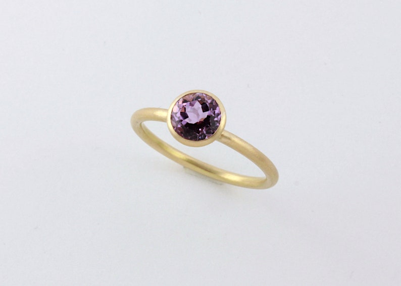 May include: A delicate gold ring with a round, bezel-set purple gemstone.