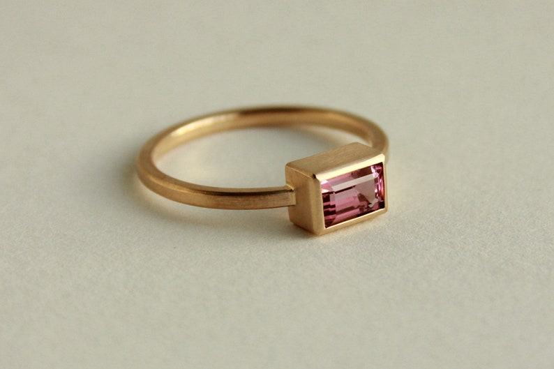 May include: A gold ring with a pink rectangular gemstone set in a gold bezel.