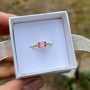 May include: A delicate gold ring with a rectangular pink gemstone set in the center. The ring is displayed in a white box.