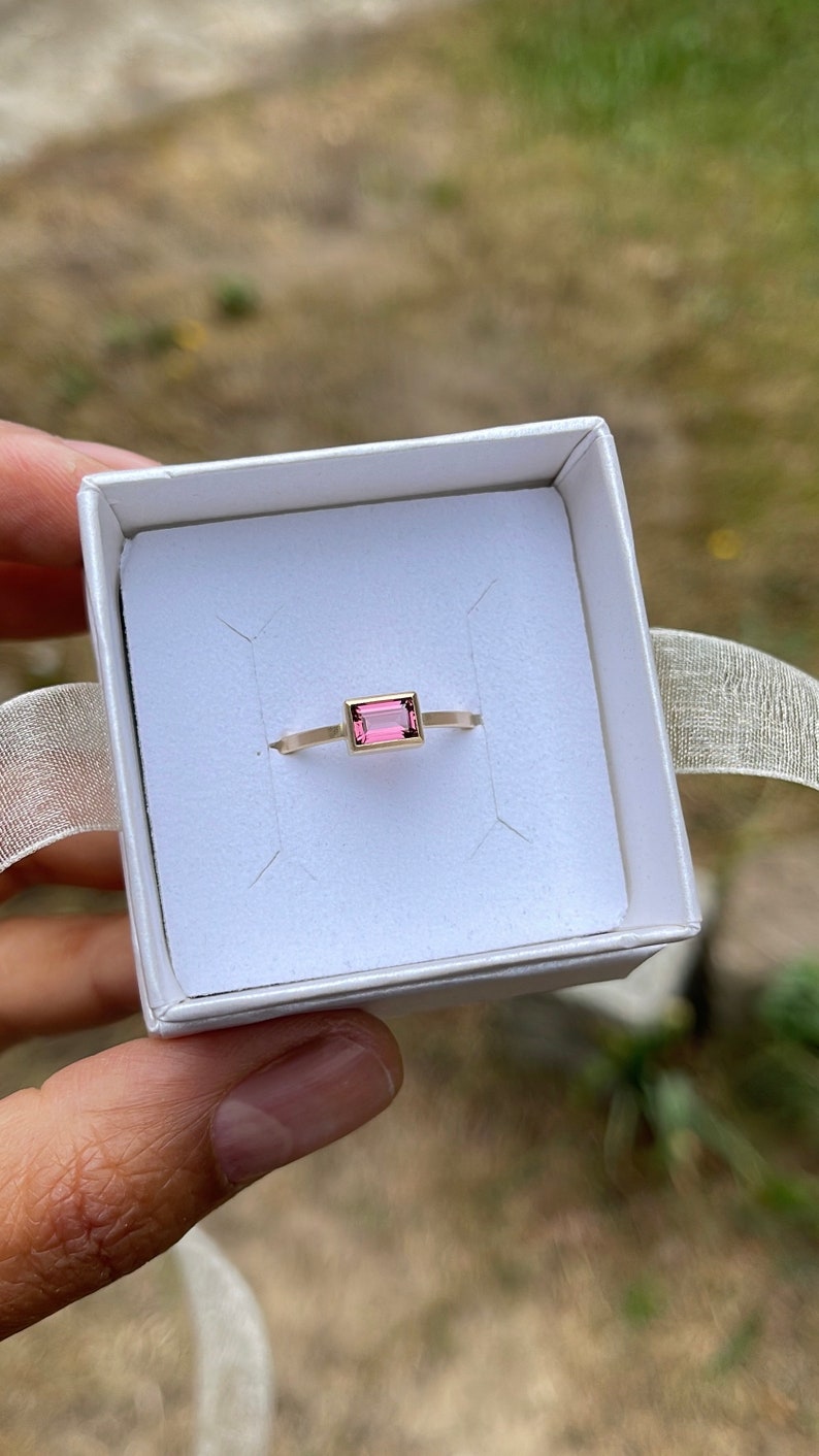 May include: A delicate gold ring with a rectangular pink gemstone set in the center. The ring is displayed in a white box.