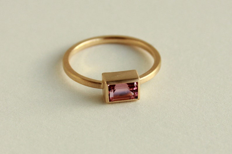May include: A gold ring with a pink rectangular gemstone set in a gold bezel.