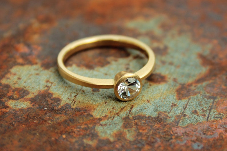 May include: A simple gold band ring with a single round clear gemstone set in a bezel setting.