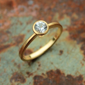 May include: A simple gold band ring with a single round clear gemstone set in a bezel setting.