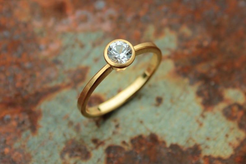 May include: A simple gold band ring with a single round clear gemstone set in a bezel setting.