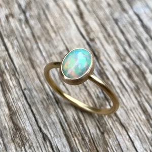 Pu&ograve; includere: Un delicato anello d'oro con una pietra preziosa di opale bianco. L'anello &egrave; posto su una superficie di legno.