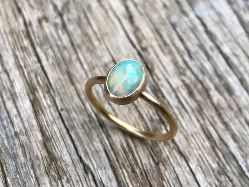 Pu&ograve; includere: Un delicato anello d'oro con una pietra preziosa di opale bianco. L'anello &egrave; posto su una superficie di legno.
